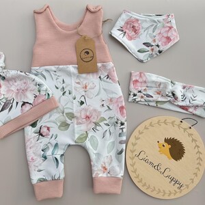 May include: A baby clothing set featuring a pink and white floral print. The set includes a jumpsuit, hat, bib, and headband. A wooden sign with a hedgehog illustration and the text "Liam & Luppy" is also included.