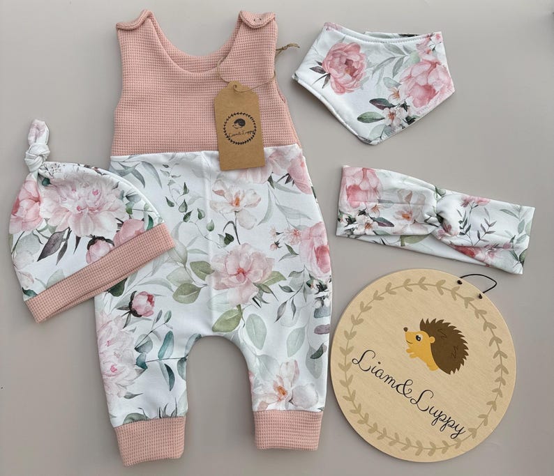 May include: A baby clothing set featuring a pink and white floral print. The set includes a jumpsuit, hat, bib, and headband. A wooden sign with a hedgehog illustration and the text "Liam & Luppy" is also included.