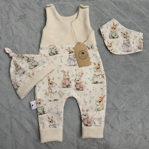 Puede incluir: Un conjunto de tres piezas para bebé que incluye un pelele color crema, un gorro a juego y un babero. La parte inferior del pelele y el gorro están estampados con conejitos con vestidos y suéteres. El babero también tiene el estampado de conejitos.