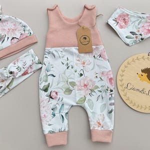 May include: A baby clothing set featuring a pink and white floral print. The set includes a romper, hat, headband, bib, and a wooden sign with a hedgehog illustration and the text "Liam & Luppy".