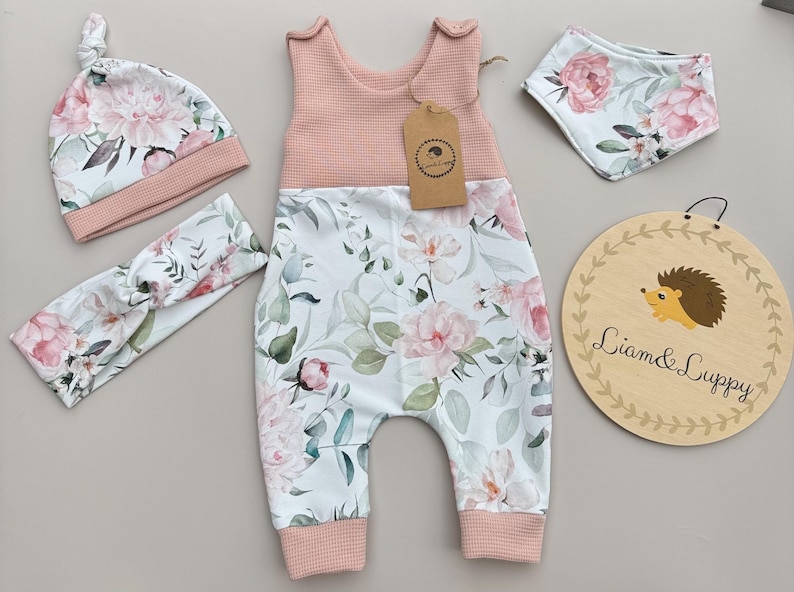 May include: A baby clothing set featuring a pink and white floral print. The set includes a romper, hat, headband, bib, and a wooden sign with a hedgehog illustration and the text "Liam & Luppy".
