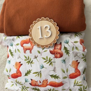Puede incluir: Una composici&oacute;n que presenta una tela de punto de gofre doblada de color &oacute;xido, un n&uacute;mero 13 de madera y una funda de almohada con un estampado de zorros y flores. La funda de almohada tiene un fondo blanco con zorros naranjas, hojas verdes y flores azules.