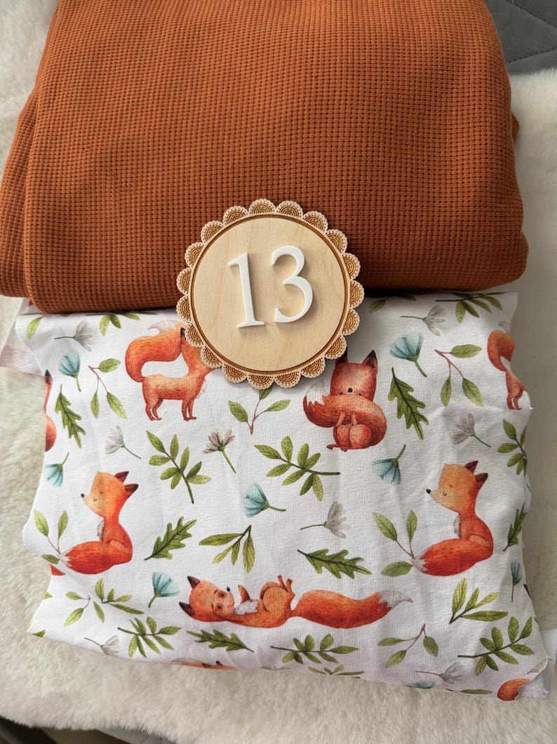 Puede incluir: Una composici&oacute;n que presenta una tela de punto de gofre doblada de color &oacute;xido, un n&uacute;mero 13 de madera y una funda de almohada con un estampado de zorros y flores. La funda de almohada tiene un fondo blanco con zorros naranjas, hojas verdes y flores azules.