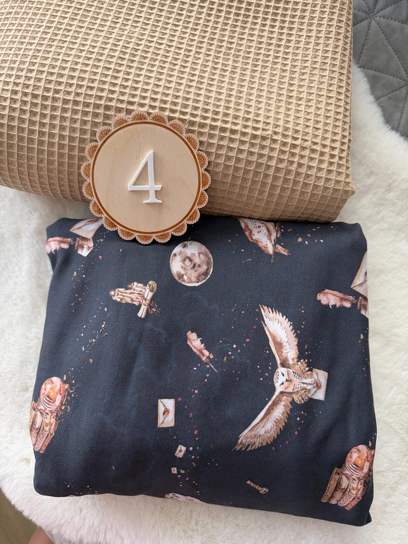 Puede incluir: Una tela azul oscuro con un estampado celestial de b&uacute;hos y astronautas, un n&uacute;mero 4 de madera y una manta con textura de gofre. La tela presenta b&uacute;hos, astronautas, plumas y sobres. El n&uacute;mero 4 es blanco sobre un c&iacute;rculo de madera.