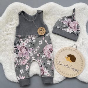 Puede incluir: Un conjunto de pelele y gorro para bebé gris con un estampado floral en rosa y blanco. El pelele tiene una parte superior de punto de gofre y el gorro tiene una parte superior anudada. También se incluye un letrero de madera con una ilustración de erizo y el texto "Liam & Luppy".