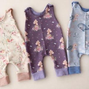 Puede incluir: Tres peleles de bebé. Uno es crema con un estampado floral, el segundo es morado con un estampado de jirafas y el tercero es azul claro con un estampado de animales marinos. Todos tienen puños y cuellos de canalé.