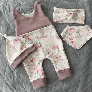 Puede incluir: Un conjunto de ropa de bebé con un diseño floral de unicornios en rosa y blanco. El conjunto incluye un pelele, un gorro, una diadema y un babero. El pelele y el gorro tienen una parte superior rosa texturizada.