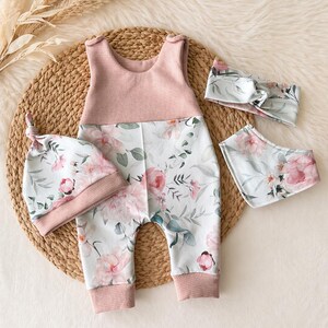 May include: A baby clothing set featuring a pink and floral print romper, a matching knotted hat, a bib, and a headband. The romper has a pink waffle-knit bodice and floral print pants. The set is designed for infants.