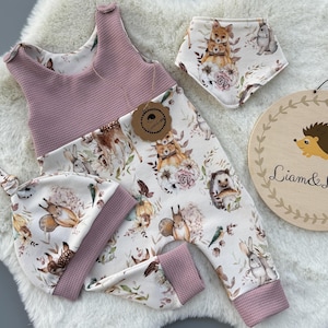 Puede incluir: Un conjunto de ropa de bebé con un pelele, un babero y un gorro, todos estampados con animales del bosque. El pelele y el gorro tienen una parte superior y un ribete de punto de gofre malva. También se incluye un letrero de madera con una ilustración de erizo y el texto "Liam & Luppy".