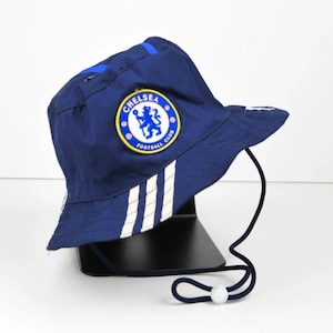 May include: A navy blue bucket hat featuring the Chelsea Football Club logo on the front. The hat has white stripes and a chin strap with a toggle. The hat is designed with a brim for sun protection.