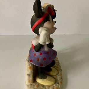 Może przedstawiać: Figurka Myszki Minnie, z czarną głową, czerwoną opaską i małym ż&oacute;łtym kapeluszem. Ma na sobie fioletową sukienkę w kropki, białe rękawiczki i ż&oacute;łte buty. Figurka stoi na teksturowanej podstawie.