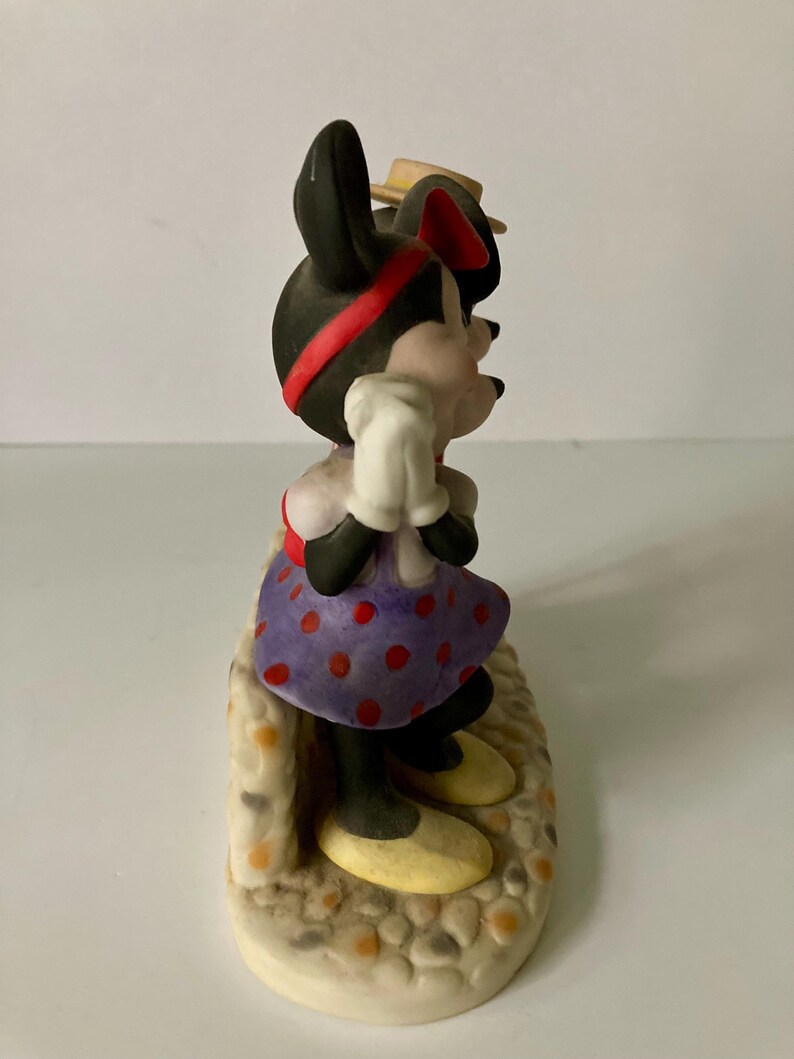 Może przedstawiać: Figurka Myszki Minnie, z czarną głową, czerwoną opaską i małym ż&oacute;łtym kapeluszem. Ma na sobie fioletową sukienkę w kropki, białe rękawiczki i ż&oacute;łte buty. Figurka stoi na teksturowanej podstawie.