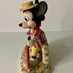 Może przedstawiać: Ceramiczna figurka Myszki Miki w czerwono-białej kurtce w paski, ż&oacute;łtych spodniach i beżowym kapeluszu. Trzyma czerwoną laskę i stoi na teksturowanej podstawie z kolorowymi plamkami. Zabytkowy przedmiot kolekcjonerski Disneya.