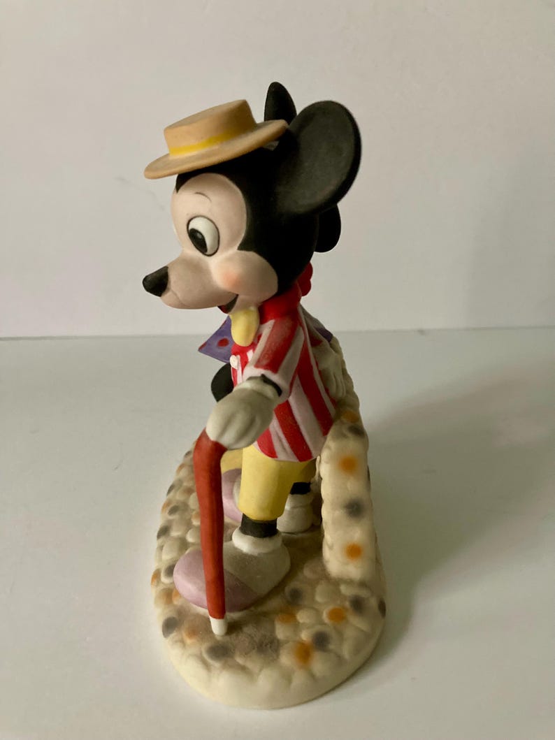 Może przedstawiać: Ceramiczna figurka Myszki Miki w czerwono-białej kurtce w paski, ż&oacute;łtych spodniach i beżowym kapeluszu. Trzyma czerwoną laskę i stoi na teksturowanej podstawie z kolorowymi plamkami. Zabytkowy przedmiot kolekcjonerski Disneya.