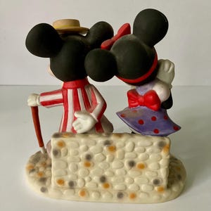 Może przedstawiać: Ceramiczna figurka przedstawiająca Myszki Miki i Minnie. Miki ma na sobie czerwono-biały pasiasty str&oacute;j i słomkowy kapelusz, trzymając laskę. Myszka Minnie ma fioletową sukienkę w kropki i czerwoną kokardę. Stoją na kamiennej podstawie.