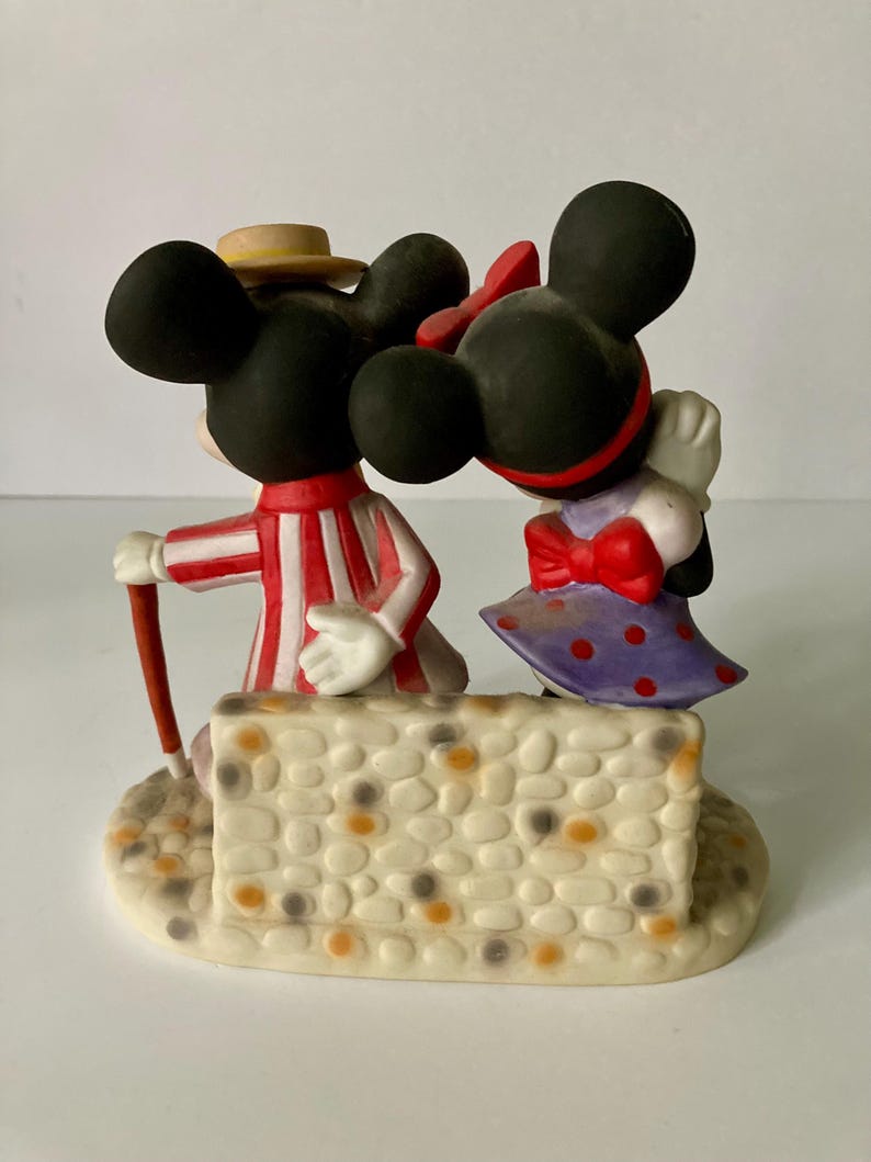 Może przedstawiać: Ceramiczna figurka przedstawiająca Myszki Miki i Minnie. Miki ma na sobie czerwono-biały pasiasty str&oacute;j i słomkowy kapelusz, trzymając laskę. Myszka Minnie ma fioletową sukienkę w kropki i czerwoną kokardę. Stoją na kamiennej podstawie.