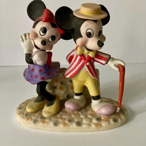 Może przedstawiać: Ceramiczna figurka Myszki Miki i Myszki Minnie. Minnie ma czerwoną kokardę, fioletową sp&oacute;dnicę w kropki i ż&oacute;łte buty. Miki ma pasiastą kurtkę, ż&oacute;łte spodnie i słomkowy kapelusz. Stoją na teksturowanej podstawie.