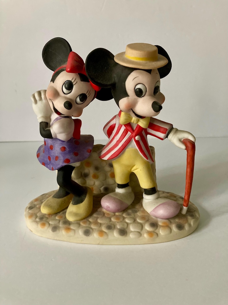 Może przedstawiać: Ceramiczna figurka Myszki Miki i Myszki Minnie. Minnie ma czerwoną kokardę, fioletową sp&oacute;dnicę w kropki i ż&oacute;łte buty. Miki ma pasiastą kurtkę, ż&oacute;łte spodnie i słomkowy kapelusz. Stoją na teksturowanej podstawie.
