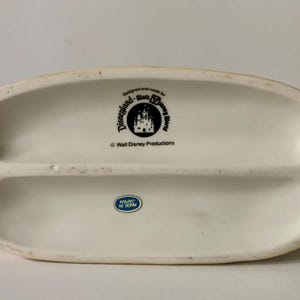 Może przedstawiać: Kremowa ceramiczna podstawa z logo Disneyland i Walt Disney World oraz napisem "Walt Disney Productions". Niebiesko-biała naklejka głosi "Made in Japan".