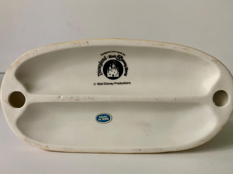 Może przedstawiać: Kremowa ceramiczna podstawa z logo Disneyland i Walt Disney World oraz napisem "Walt Disney Productions". Niebiesko-biała naklejka głosi "Made in Japan".