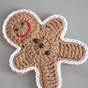 May include: A crocheted gingerbread man coaster with a brown body, white trim, and black bead eyes. The gingerbread man has a red stitched smile. This handmade coaster is perfect for the holiday season.