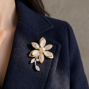 Puede incluir: Un broche de flor en tono dorado con cinco pétalos que presentan inserciones blancas iridiscentes, una perla redonda central y pequeños cristales transparentes incrustados a lo largo de los bordes y en la extensión inferior.