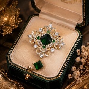 May include: A gold-toned brooch featuring a large square-cut dark green center stone, surrounded by smaller clear white crystals and tiny pearls. The ornate metal frame includes intricate scrollwork and a dangling square green stone pendant.