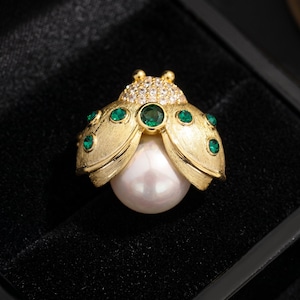 May include: Gold-toned ladybug brooch with a large white pearl body. The brooch features emerald green accents and a head encrusted with small, clear crystals. The wings have a textured finish, and the brooch is set against a black background.