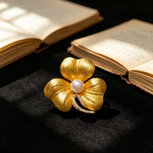 May include: A gold-colored flower-shaped brooch with a pearl center and a curved accent of small, sparkling stones. The brooch is set against a dark background, with open books in the background.
