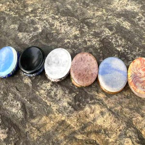 May include: Six oval-shaped stone plugs in a row, each with a unique color and pattern. The colors include blue, black, white, brown, and a mix of blue and orange. The plugs are set on a textured, gray-brown stone surface.