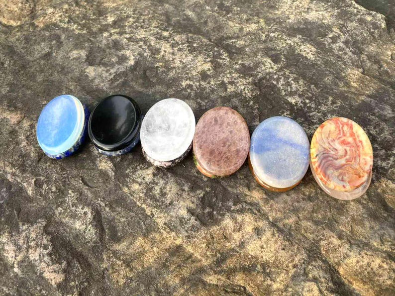 Puede incluir: Seis tapones de piedra ovalados en fila, cada uno con un color y patr&oacute;n &uacute;nicos. Los colores incluyen azul, negro, blanco, marr&oacute;n y una mezcla de azul y naranja. Los tapones est&aacute;n colocados sobre una superficie de piedra texturizada gris-marr&oacute;n.