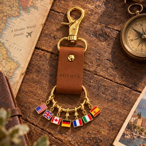 May include: A gold-toned metal keychain featuring a brown leather strap embossed with the word ROAMER. The strap is attached to a gold lobster clasp and a circular ring holding seven small, square, enamel-coated charms depicting national flags.