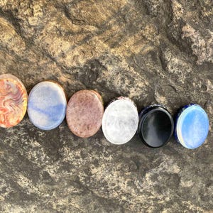 Puede incluir: Una colecci&oacute;n de siete piedras ovaladas en varios colores y patrones. Las piedras incluyen tonos de rojo, azul, marr&oacute;n, blanco y negro, con una &uacute;ltima piedra en azul y blanco. Las piedras est&aacute;n dispuestas en fila.
