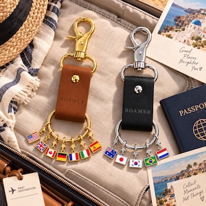 May include: Two leather keychains with metal hardware, one in brown with gold-tone accents and one in black with silver-tone accents. Each features the word ROAMER embossed on the leather strap and a ring adorned with multiple small enamel flag charms.
