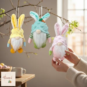 May include: Three Easter gnome ornaments with bunny ears suspended from a tree branch. The gnomes are yellow, blue, and pink, with white beards and colourful outfits. A table with a mug and a basket of Easter eggs is also visible.
