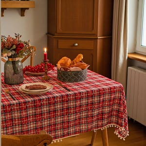 Puede incluir: Un mantel a cuadros rojos y blancos con un ribete de pompón blanco cubre una mesa de madera. Una cesta de pan, un jarrón de flores y un plato de comida están sobre la mesa. Una vela encendida está en un candelabro de latón.