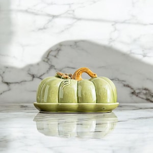 Puede incluir: Un plato de cerámica en forma de calabaza, de color verde claro, con tapa y tallo marrón. Ideal para servir o decorar. La calabaza tiene estrías verticales y descansa sobre una base a juego.