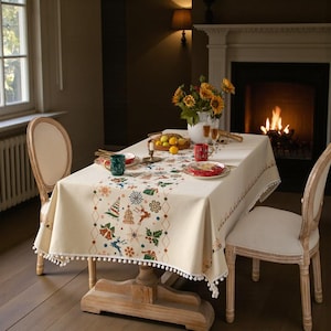 May include: A cream-coloured rectangular tablecloth with a Christmas-themed pattern featuring trees, snowflakes, and reindeer. The tablecloth has a decorative border and white pom-pom trim. The table is set with plates, mugs, and a vase of sunflowers.