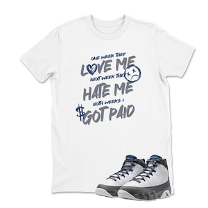 May include: White t-shirt with the text "ONE WEEK THEY LOVE ME, NEXT WEEK THEY HATE ME, BOTH WEEKS I GOT PAID" in blue and gray. A pair of gray, white, and blue sneakers are at the bottom.