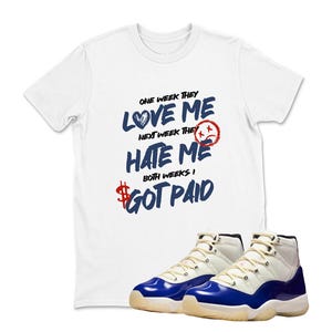 May include: White t-shirt with the text "ONE WEEK THEY LOVE ME, NEXT WEEK THEY HATE ME, BOTH WEEKS I GOT PAID" in blue and red. The shirt is paired with white and blue sneakers.