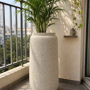 May include: A tall, cylindrical floor planter with a textured, speckled off-white finish. The planter has a rounded base and a slightly tapered top opening, containing a green palm plant with long, slender, feathery fronds extending upwards.