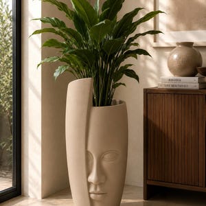 Puede incluir: Una maceta alta de color beige con un diseño de rostro esculpido, que contiene una exuberante planta verde. La maceta tiene una estética moderna y minimalista. Las hojas grandes y vibrantes de la planta contrastan con los tonos neutros de la maceta y la decoración.