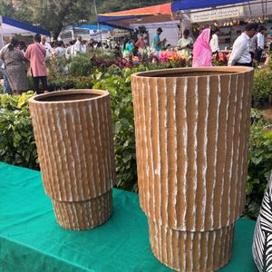 Peut inclure: Deux jardinières cylindriques avec une surface beige texturée présentant des rainures verticales blanches. Les jardinières ont un design étagé avec une section supérieure plus large et une base plus étroite. Elles sont posées sur une surface verte.