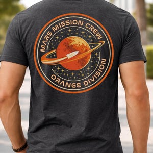 May include: Dark grey t-shirt with a circular graphic. The graphic features the text "Mars Mission Crew Orange Division" around an orange planet with a white rocket and an orange ring. The design is outlined in orange and white.