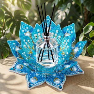 May include: A clear glass reed diffuser bottle with four black reeds sits in the center of a decorative blue lotus flower base. The base features textured blue petals adorned with white faux pearls and small gold accents in a layered design.
