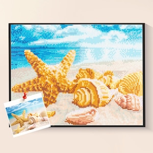 May include: A framed diamond painting depicts a beach scene with a large starfish, seashells, and a sandy shore under a blue sky. The artwork is in a pixelated style, with a small photo pinned to the frame.