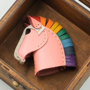 May include: A pink leather unicorn head with a rainbow mane, displayed in a wooden box. The unicorn's head is light pink with a white and tan face. The mane features red, orange, yellow, green, blue, indigo, and purple segments.