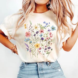 May include: Cream-colored t-shirt featuring a vibrant watercolor floral design. The design includes a variety of colorful flowers, including pink, blue, yellow, and purple blooms, with green foliage. The shirt is paired with blue denim jeans.