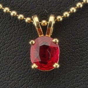 May include: An oval-cut deep red gemstone pendant set in a four-prong gold mounting, suspended from a gold-toned ball chain necklace. The faceted stone displays a vibrant crimson hue, securely held by the polished gold metal prongs and bail.