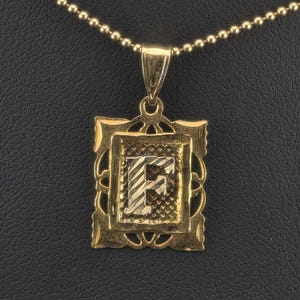 May include: A gold-toned pendant featuring the letter F in a textured, rectangular frame with decorative edges, suspended from a matching gold-toned ball chain necklace. The letter F has a striped finish and is centered within the ornate border.